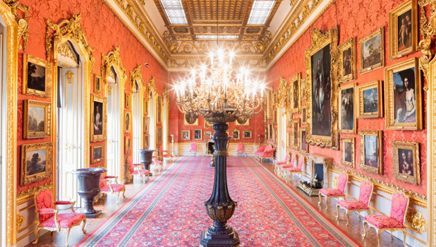 Ornate gallery room with red and gold décor, upholstered chairs, gold-framed paintings, a large chandelier and tall windows with gold trim frames.